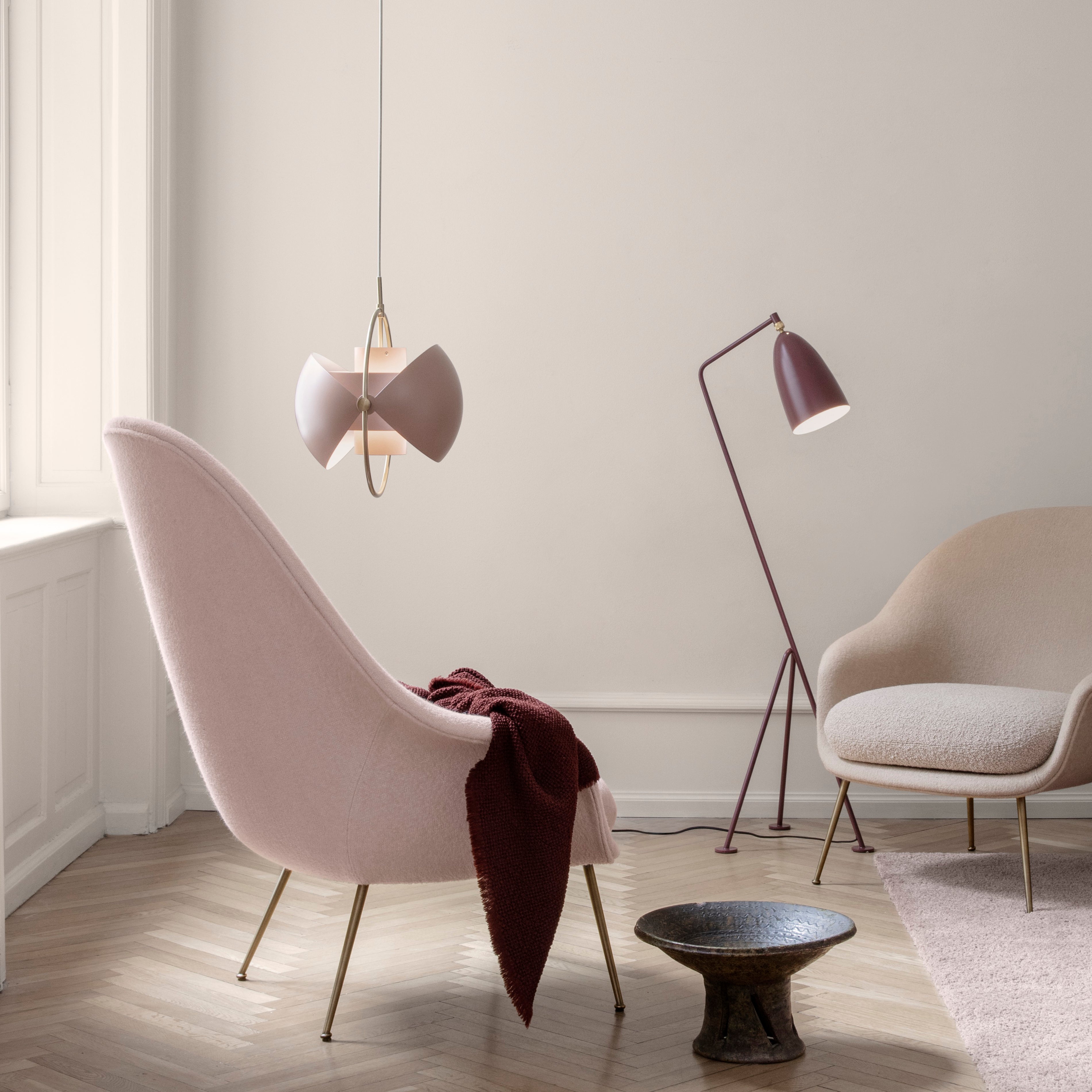 A pink pendant with two fan-shaped shades and a brass ring hangs above a pink armchair in a pale room