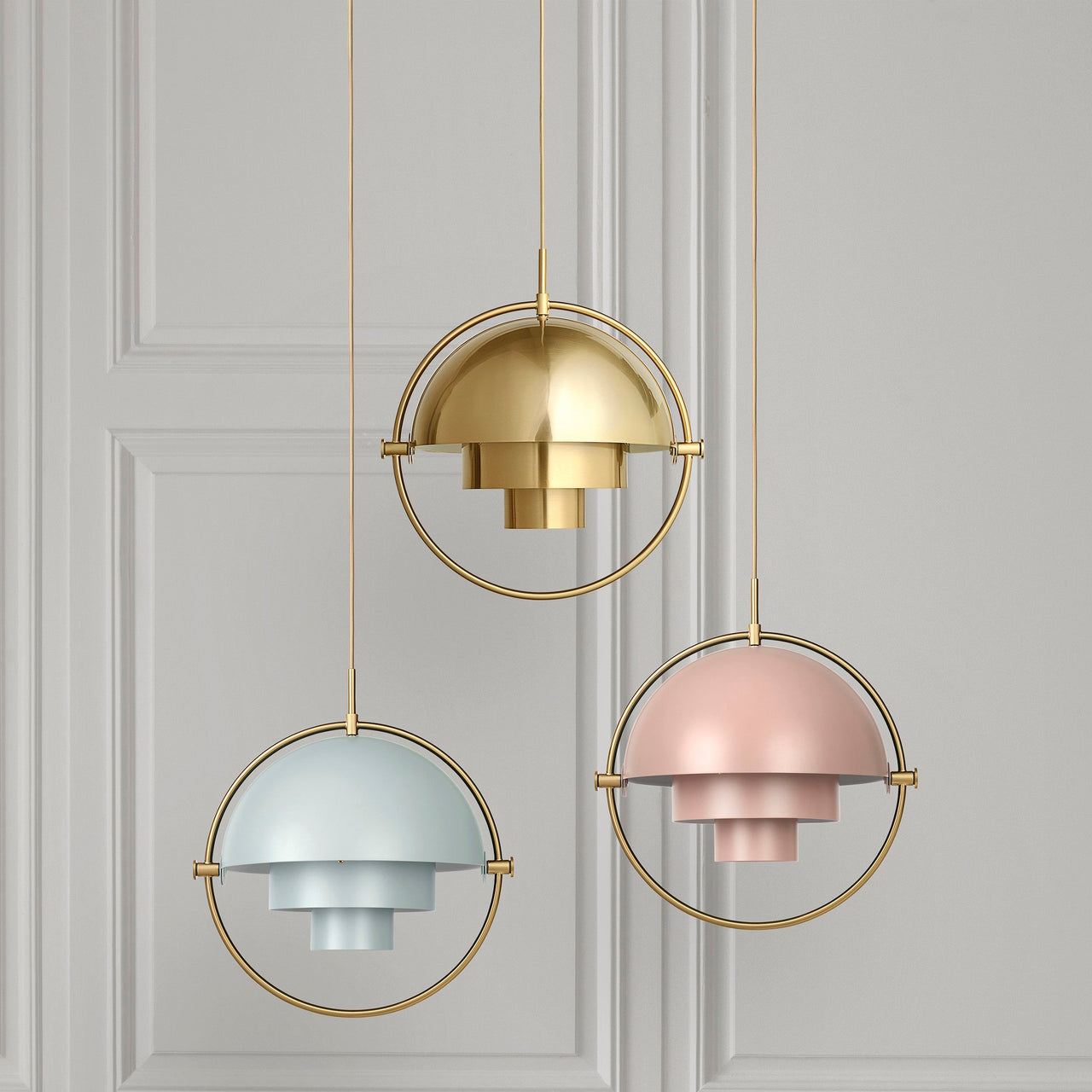 Three domed pendant lights in brass, pale blue and soft pink, each in a thin brass ring, hang before a panelled grey wall