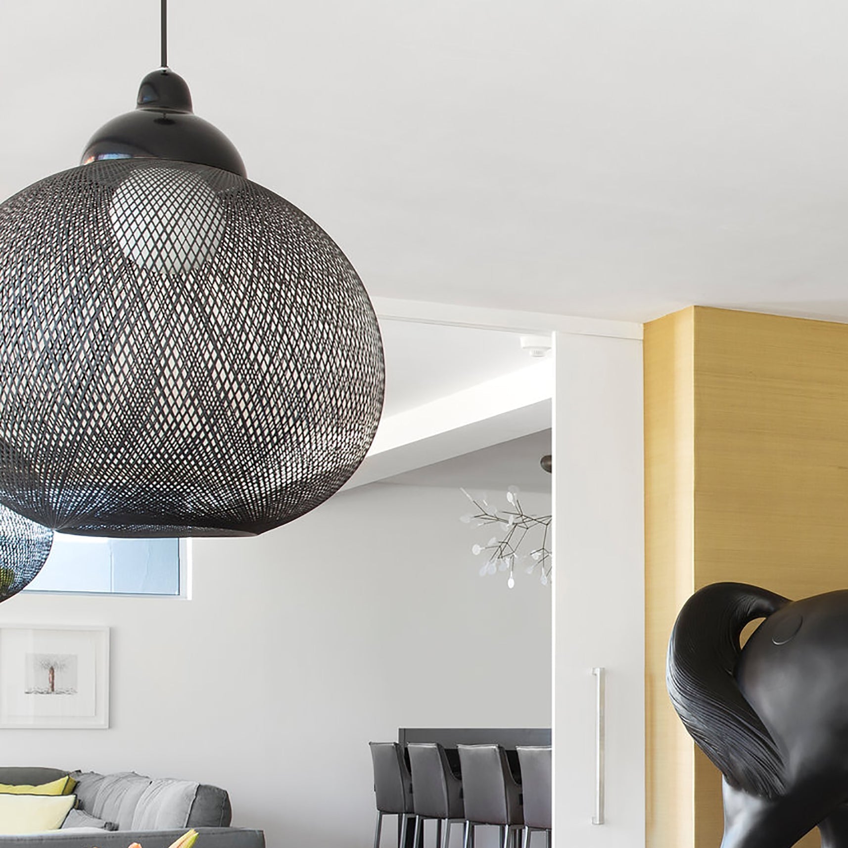 A large black woven mesh pendant lamp hangs in a bright room with a grey sofa and a black sculpture