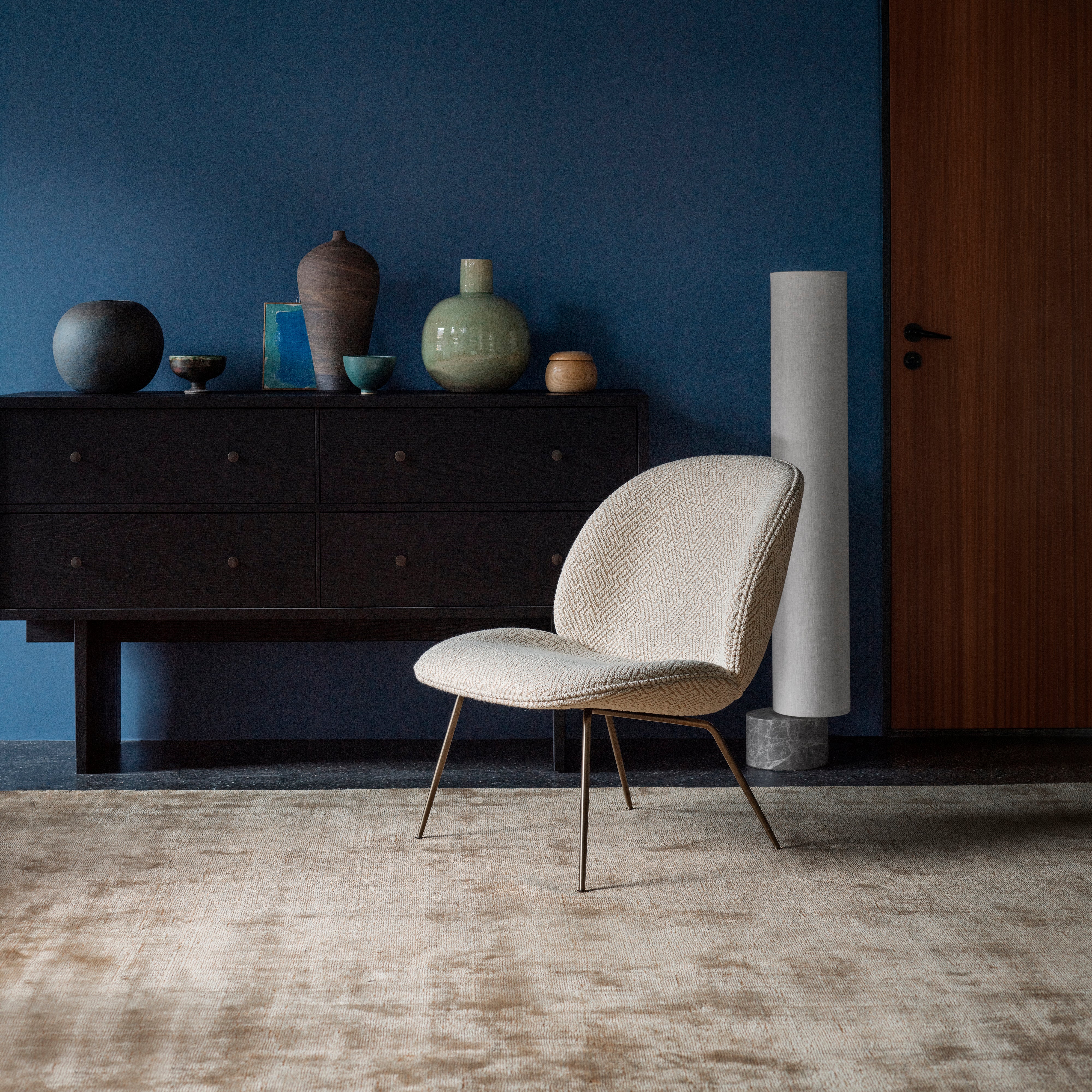 A dark wood sideboard with six drawers against a deep blue wall, with a cream patterned chair in front