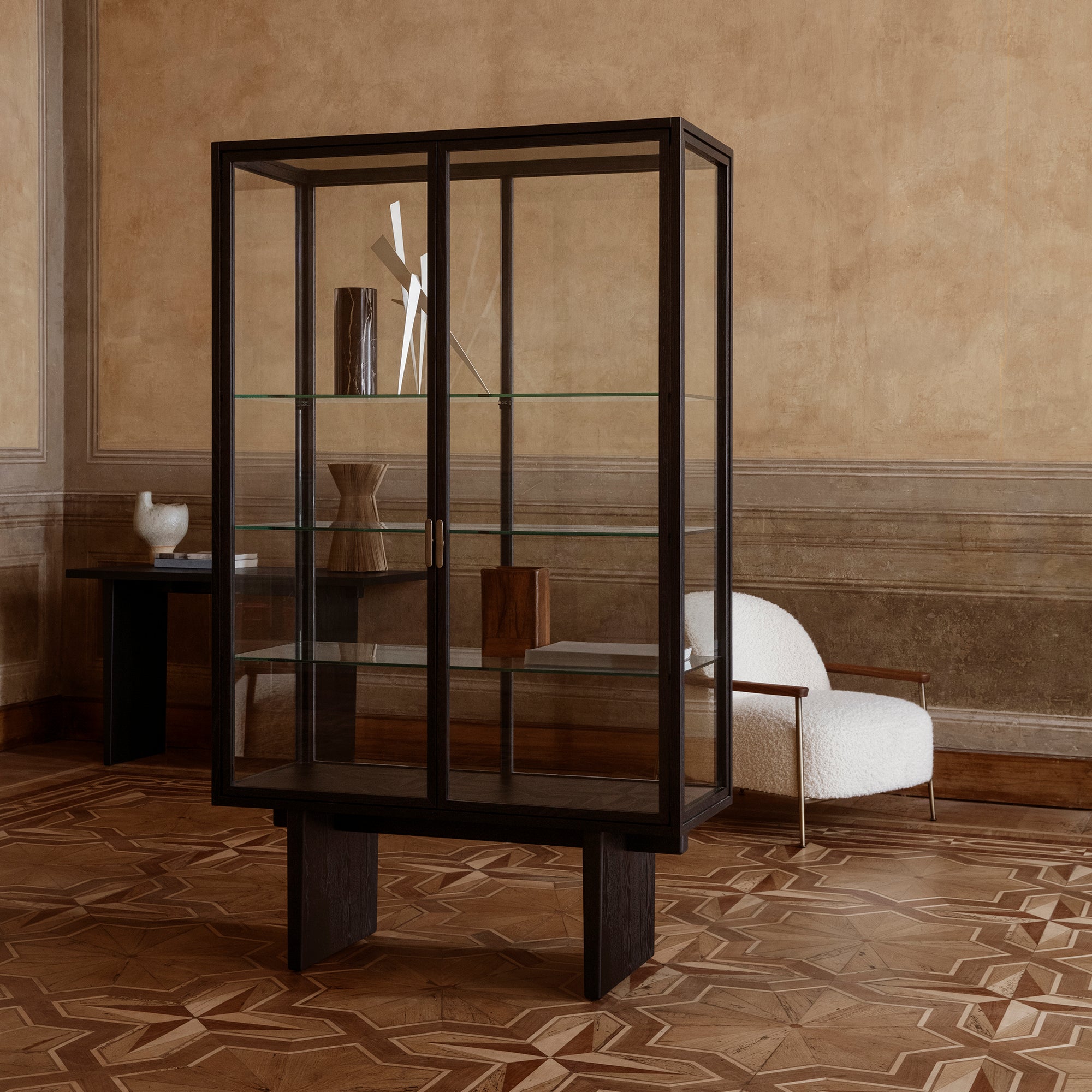 A tall glass-fronted cabinet with a dark wood frame and glass shelves in a room with patterned parquet floor