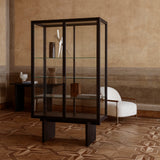 A tall glass-fronted cabinet with a dark wood frame and glass shelves in a room with patterned parquet floor