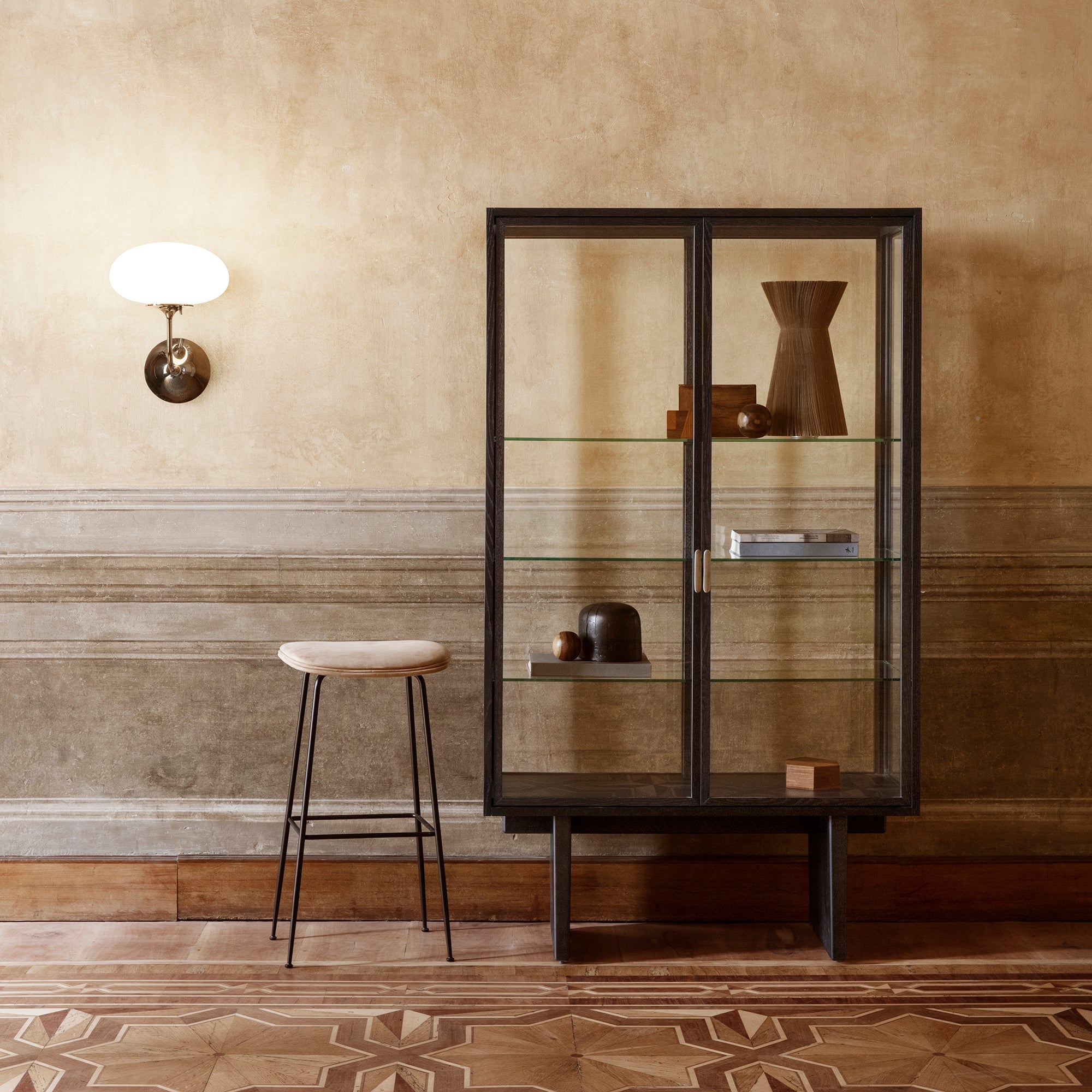 A tall glass-fronted cabinet with a dark wood frame and shelves, beside a bar stool and wall light